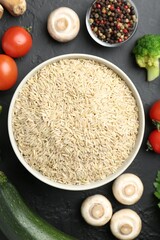 Raw brown rice in bowl among fresh products on dark textured table, flat lay