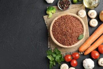 Raw brown rice in bowl among fresh products on dark textured table, flat lay. Space for text