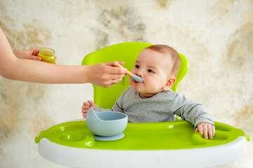 mom feeds baby with a spoon, baby food concept, little baby boy eats mashed vegetables in a green high chair