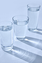 Three glasses filled with water stand on light surface, casting long, blurry shadows. Light refracts through water, creating sparkling effect. Mock up. Concept of hydration, drink, beverage. Ad