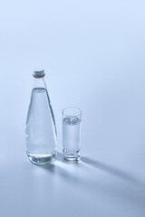 Clear glass bottle and matching glass, both filled with transparent water, on light blue surface, casting long shadows. Mock up. Concept of refreshment. Negative space for text.