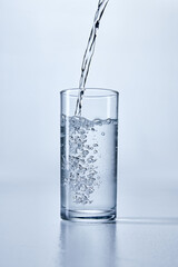 Close-up photo of transparent water being poured into a clear, cylindrical glass, showing bubbles and splashes, against soft background. Mock up. Concept of dynamic refreshment, diet.