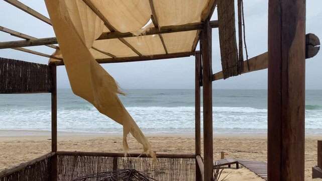 beige tattered canopy flutters in the wind, empty beach with gentle ocean waves under a cloudy sky. The muted colors, worn fabric, melancholic, nostalgic atmosphere.