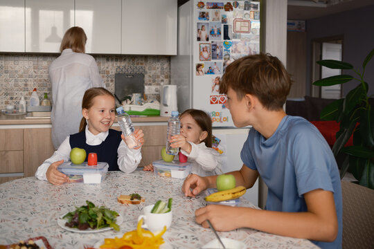 Children sit at a table packing nutritious lunches with fruits and water while getting ready for school. A parent assists in the kitchen, creating a lively morning routine atmosphere.