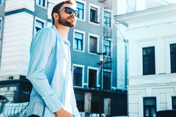 Portrait of handsome smiling stylish hipster lambersexual model.Modern man dressed in blue shirt. Fashion male posing near skyscraper on the street background. Outdoors at sunset