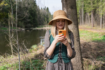 A young blonde woman in a straw hat and vintage dress is standing in a serene forest by the river,...
