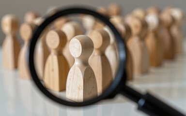 Fototapeta premium Magnifying glass focuses on a wooden figure among a group, highlighting one individual from the crowd, on a neutral surface.
