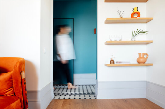 A cozy modern living room with white walls, an armchair and wooden shelves with household items. The silhouette of a walking person. - Powered by Adobe