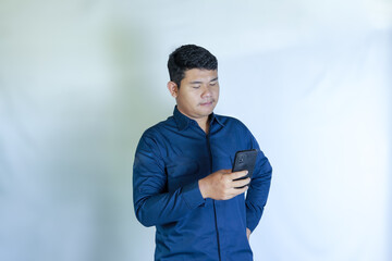 Portrait of excited Asian man in blue shirt holding mobile phone with sad expression on confused face while looking at smartphone. Isolated image on white background