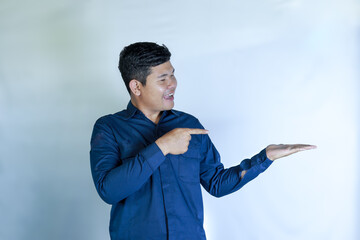 Handsome Asian Man Smiling in Blue Shirt Pointing to Empty Space in Hand with Cheerful Expression, Presenting Product, Idea or Advertising Message, Isolated on White Background
