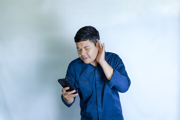 Portrait of excited Asian man in blue shirt holding mobile phone with happy expression looking at smartphone and hand next to ear. Isolated image on white background