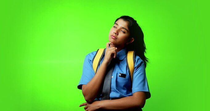 Portrait of Indian Asian teenage girl in school uniform with backpack standing against green chroma screen, looking thoughtful while placing hand on her chin, symbolizing curiosity, decision-making