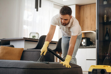 Fototapeta premium Care of sofa, using vacuum cleaner. Male professional cleaning service worker is domestic room