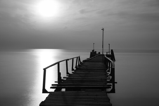 Old wooden pier extends into the sea on Koh Samet, Thailand. Crooked wooden rails are attached to the sides. Long exposure makes the sea smooth. Sunlight creates a shimmering path to the horizon