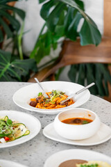 A beautifully presented dish featuring pasta with vegetables, accompanied by a bowl of soup on a textured tabletop. Lush green plants in the background add a fresh ambiance.