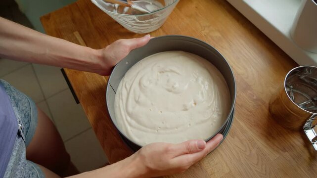 Woman holding baking pan filled with smooth cake batter, preparing for oven baking. Female placing ready batter into round pan for further cooking process. Lady setting up cake mixture in metal pan on