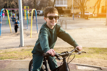 A smiling boy in sunglasses rides a bike at a sunny playground with colorful equipment. Perfect for childhood, outdoor fun, and active lifestyle themes.