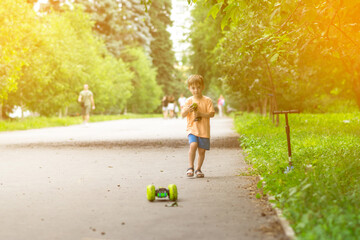 A smiling boy in summer clothes holds a green remote control while squatting on a park path. Outdoor fun with toys on a warm and sunny day.