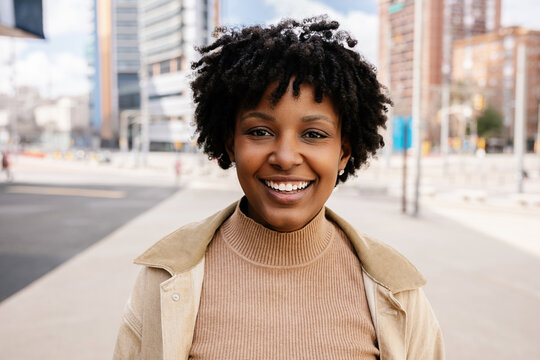 Happy young African American girl smiling at camera standing at city street. Outside portrait of joyful beautiful black woman over urban background