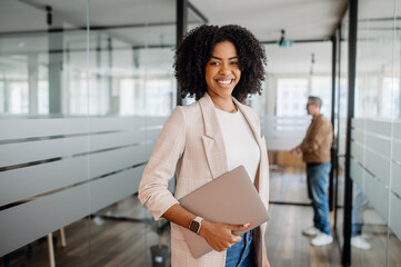Confident businesswoman standing in a modern office with a laptop, symbolizing leadership,...