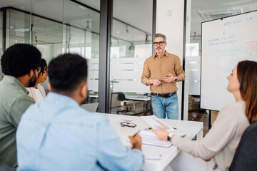 Obraz premium A professional business coach leads a strategic planning session in office. A diverse team listens attentively, taking notes and engaging in discussion. Teamwork, leadership, and professional growth.