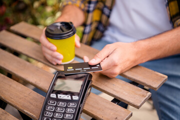 Young man making a contactless payment with a card in a cafe outdoors, enjoying a casual moment with a coffee under natural light.