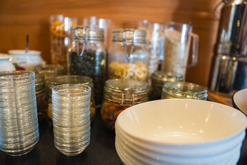 Neatly arranged glass jars with dried fruits, nuts, and cereals on a wooden surface, with glasses, bowls, and a metallic dispenser in the foreground.