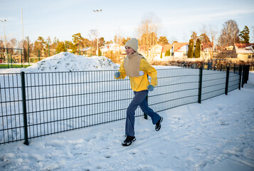 Woman in yellow jacket and hat jogging on a snowy outdoor park . Concept of winter fitness