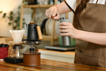 A woman is grinding coffee beans in a coffee grinder. The coffee grinder is on a wooden table next to a few other items.
