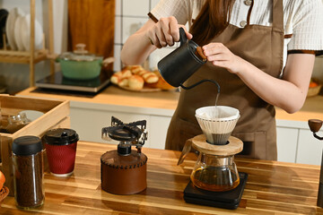 A woman is making coffee and pouring it into a cup. The scene is set in a kitchen with various items such as a scale, a cup, a bottle, and a bowl. The woman is wearing an apron.
