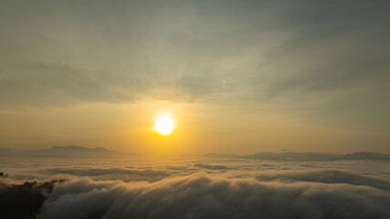 Breathtaking aerial view of the sun rising over a mountain valley covered in a dense sea of rolling clouds. The golden light illuminates the mist, creating a dramatic and ethereal atmosphere at dawn