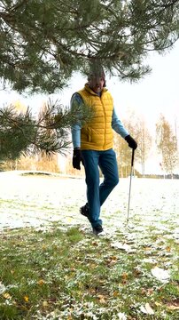 A golfer in a yellow vest and winter hat prepares to chip a ball from the rough onto a snow-covered golf course, surrounded by autumn trees.
