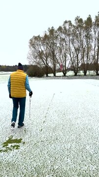 A golfer in a yellow vest and winter hat is putting on a snow-covered green near a red flag, surrounded by autumn trees.
