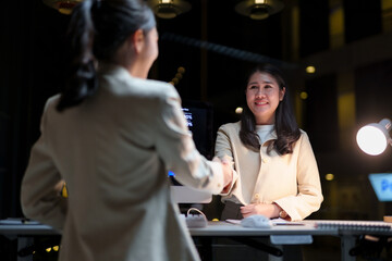 Two Asian businesswomen shaking hands in a modern office at night, celebrating the successful closure of a deal and partnership