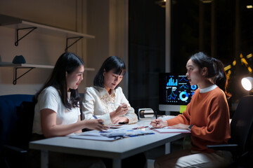 Group of young Asian businesswomen reviewing marketing data and analyzing financial charts on a computer screen late at night