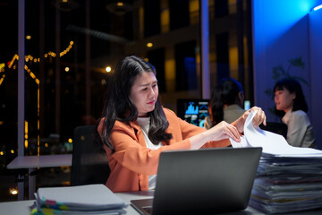 Exhausted businesswoman working overtime, laptop open, colleagues blurred behind her in dimly lit corporate workspace