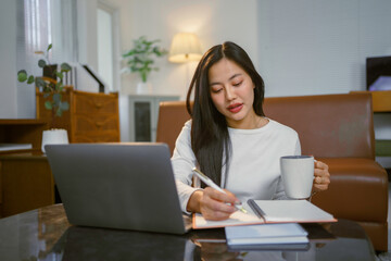 Young Asian woman working from home, drinking coffee and taking notes on a notebook while using her laptop