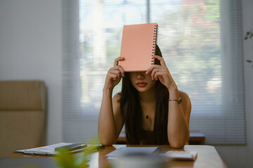 Young businesswoman is holding a notebook in front of her face while sitting at her desk in the...