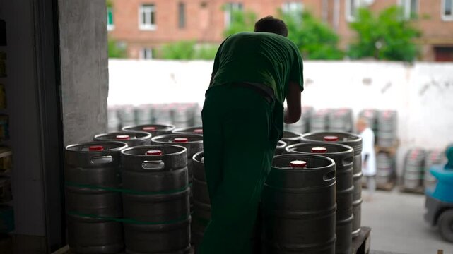 A worker seals and places security seals on metal beer kegs before shipment. The process ensures product integrity and quality control before distribution. In the background, additional kegs are stack
