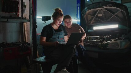 Two mechanics are focused on a laptop, collaborating to solve a car problem in a dimly lit garage. Tools are scattered around, highlighting the busy environment.