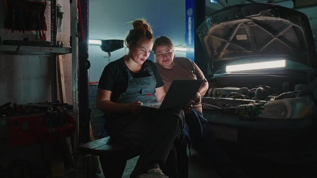 Two mechanics are focused on a laptop, collaborating to solve a car problem in a dimly lit garage. Tools are scattered around, highlighting the busy environment.