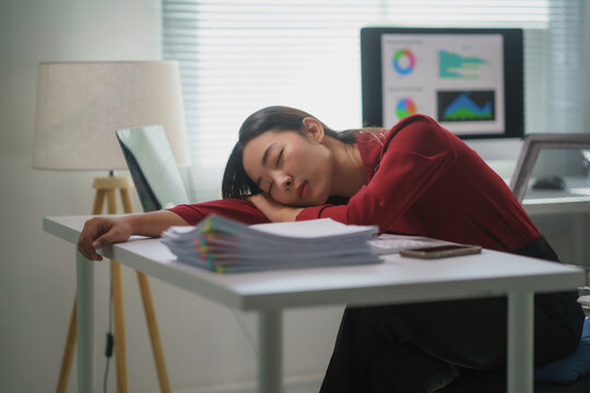 Young Asian office worker sleeping on desk at workplace after working hard on computer in modern office