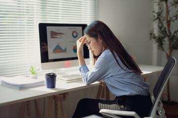 Young Asian manager suffering from headache while analyzing marketing data on desktop computer in...