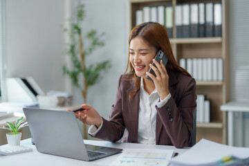 Young Asian manager working on laptop and discussing project with client on mobile phone, sitting at desk in modern office