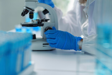 Close up of scientist's gloved hands adjusting microscope, with test tubes in foreground,...