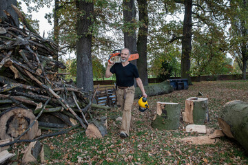 Smiling lumberjack carrying chainsaw and safety helmet in autumn forest.
