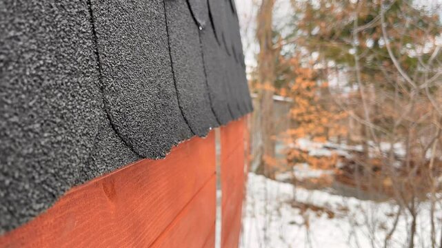 A close-up of the roof with black asphalt shingles and orange wooden siding. In the background, snow-covered trees and a forest landscape are visible.