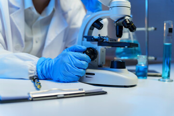 Scientist wearing gloves adjusting microscope for medical research in laboratory with test tubes and clipboard
