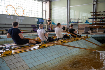 Rearview rowing male team works with paddles in pool simulator while coach observes. Sportsmen team focus on technique coordination and preparing for upcoming competitions, hobby and exercising