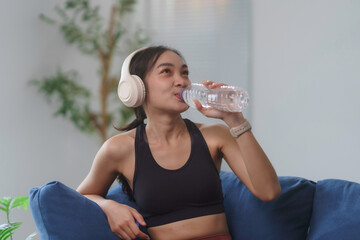 Asian athletic woman wearing headphones, enjoying a refreshing drink of water while relaxing after...